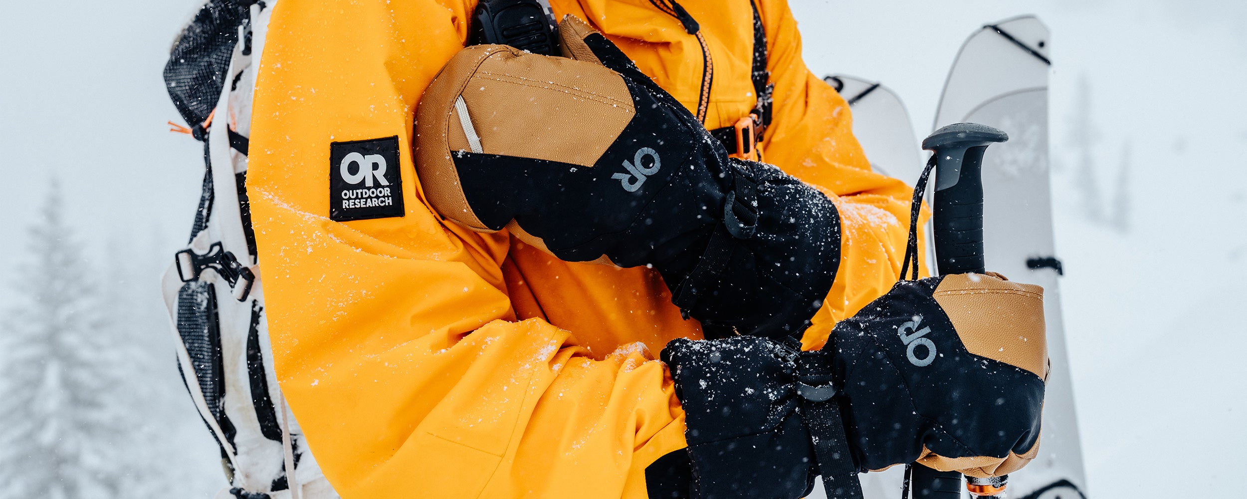 A man in the snow reaches for his walkie-talkie while wearing the Arete Modular Gore-Tex Mitts in Black/Dark Natural. 