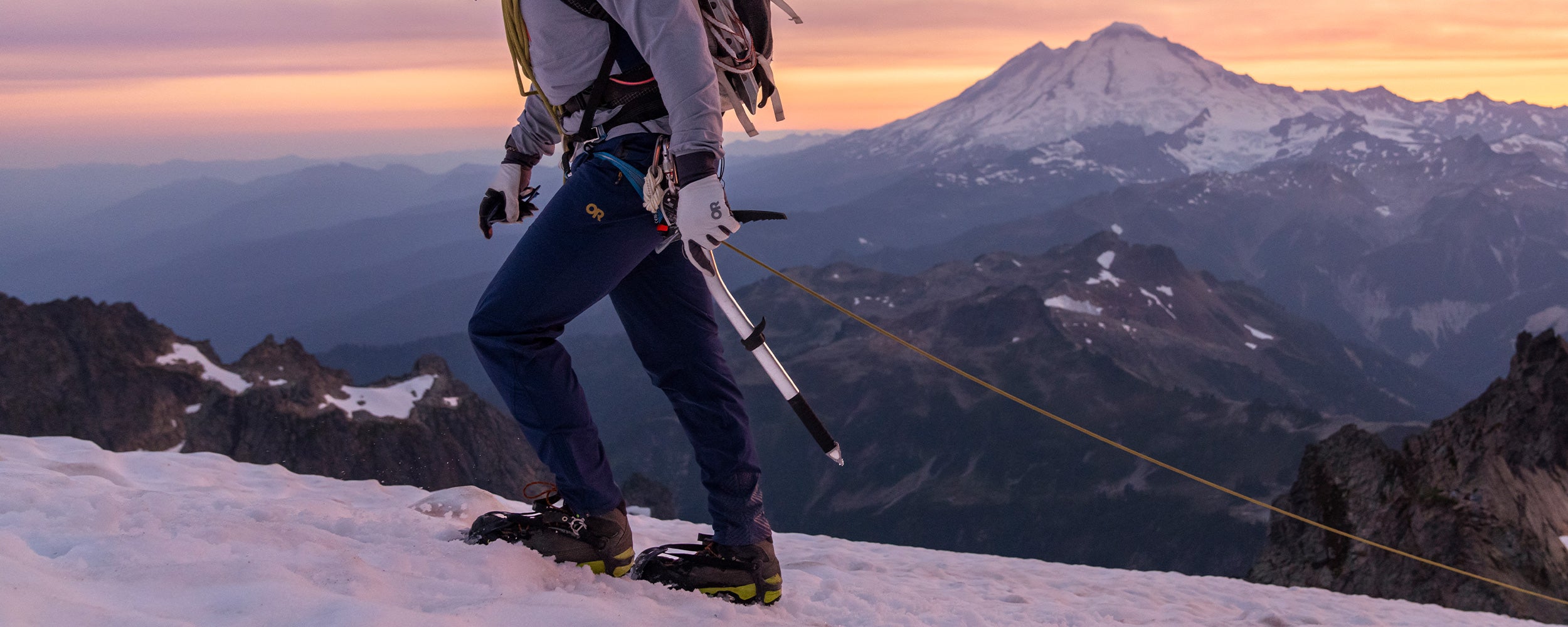 Outdoor Research athlete Max Djenohan climbs a snow-covered peak as the sun sets over the mountains in the background.