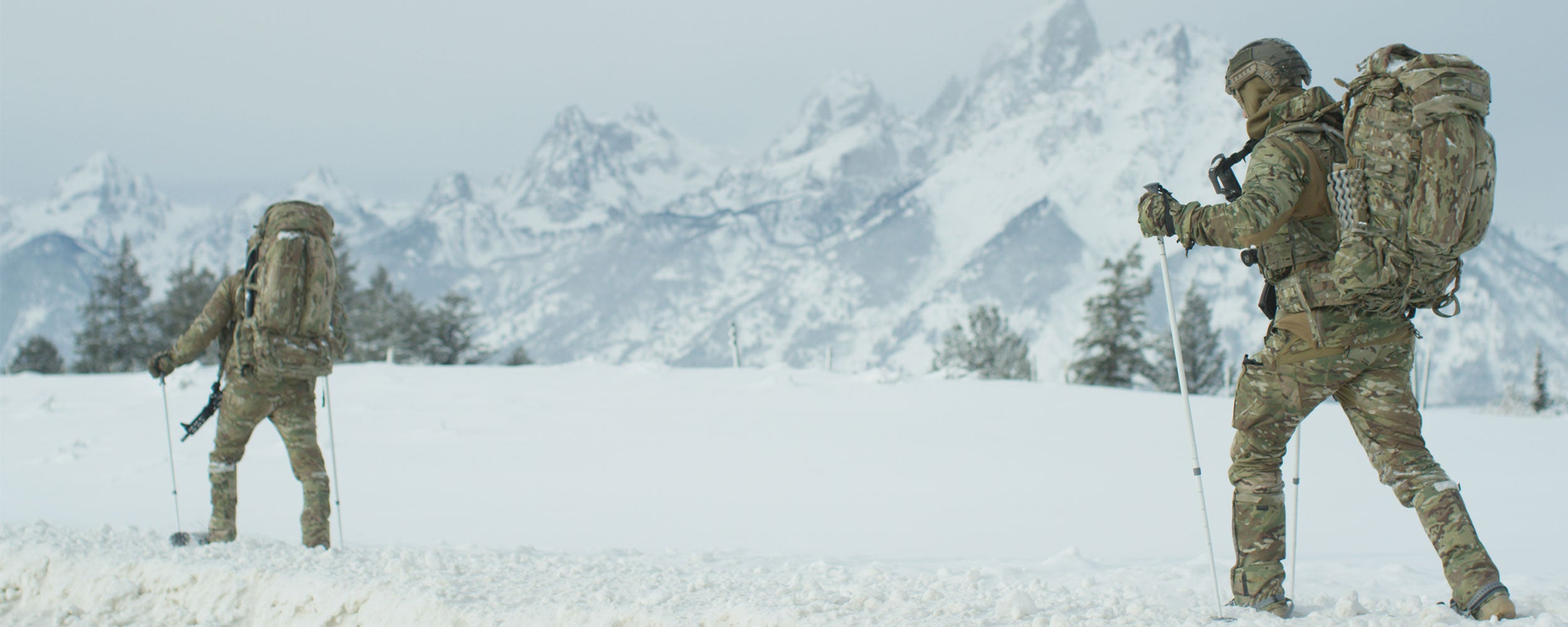 Two skiers in tactical gear cross a snowy field.