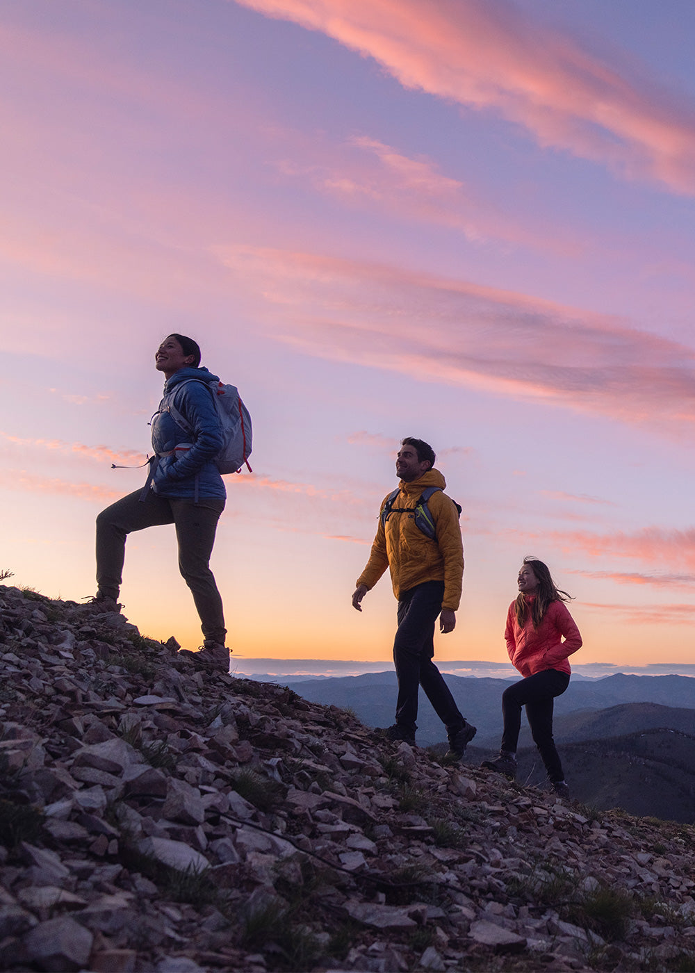 Three friends hike up a trail with the mountains and sun setting in the background.