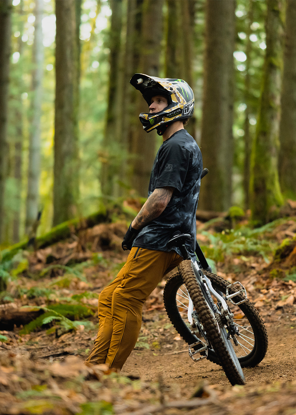 Man resting on the seat of his mountain bike wears the Freewheel Short Sleeve Jersey in Black Cloud Scape/Black. 