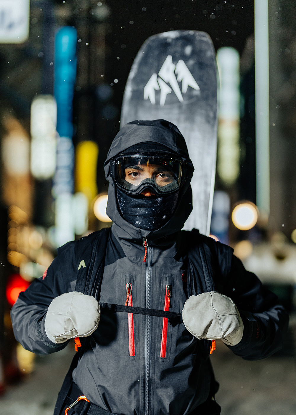 Man standing in a snowy city at night holds the straps of his ski backpack wearing a black balaclava. 