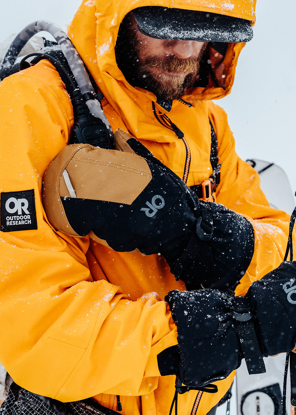 A man in the snow reaches for his walkie-talkie while wearing the Arete Modular Gore-Tex Mitts in Black/Dark Natural. 