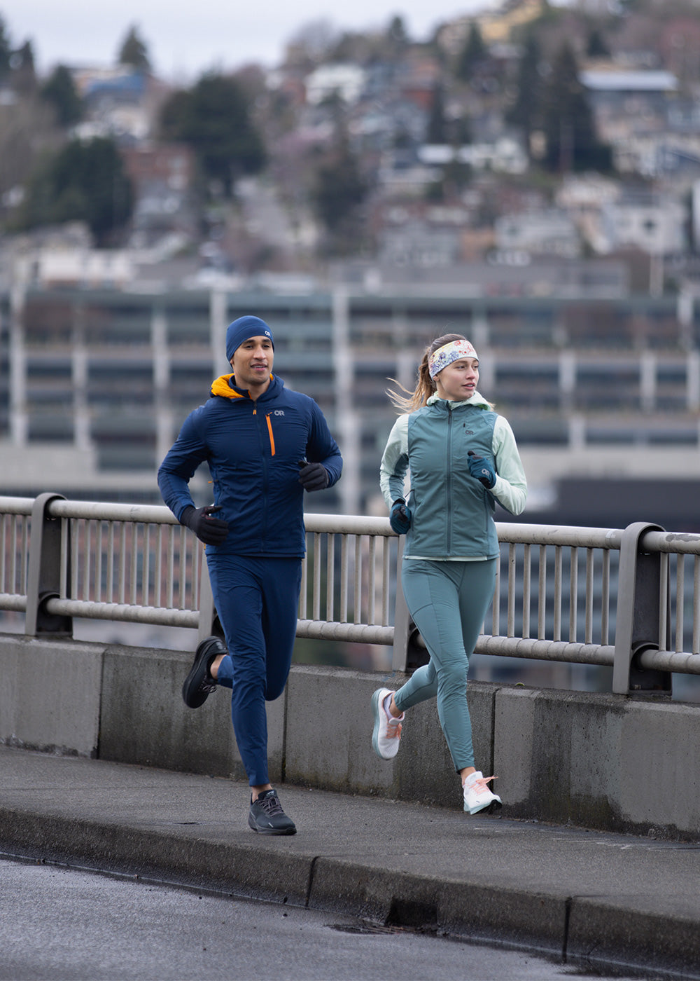 Two friends run together on a bridge while wearing the Outdoor Research Deviator Collection. 