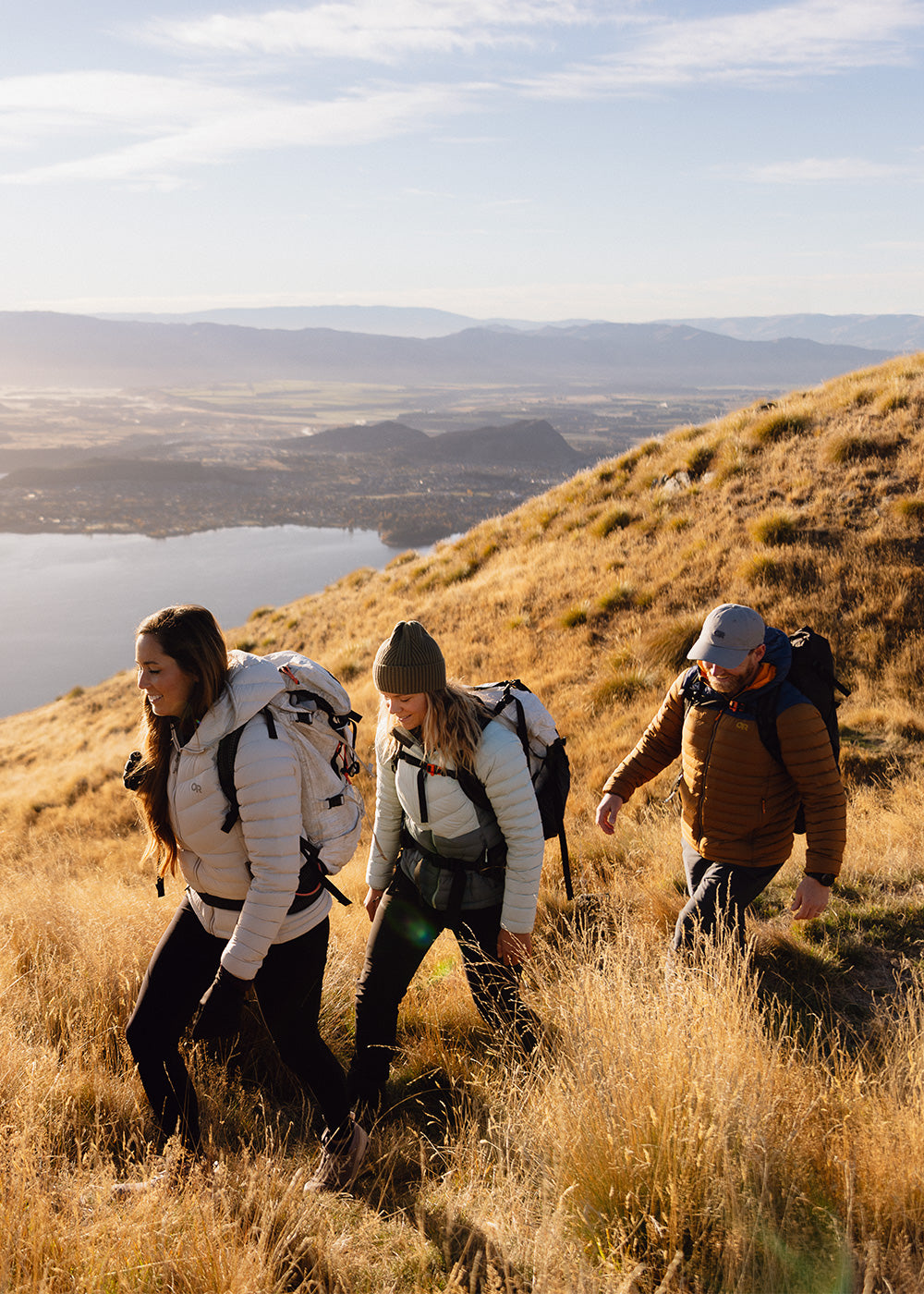 A group of friends hike along a grassy path with a lake in the background.