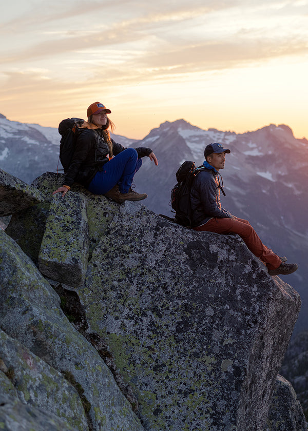 A man and a woman sit on a rocky outcropping as the sun sets behind the mountains in the background.