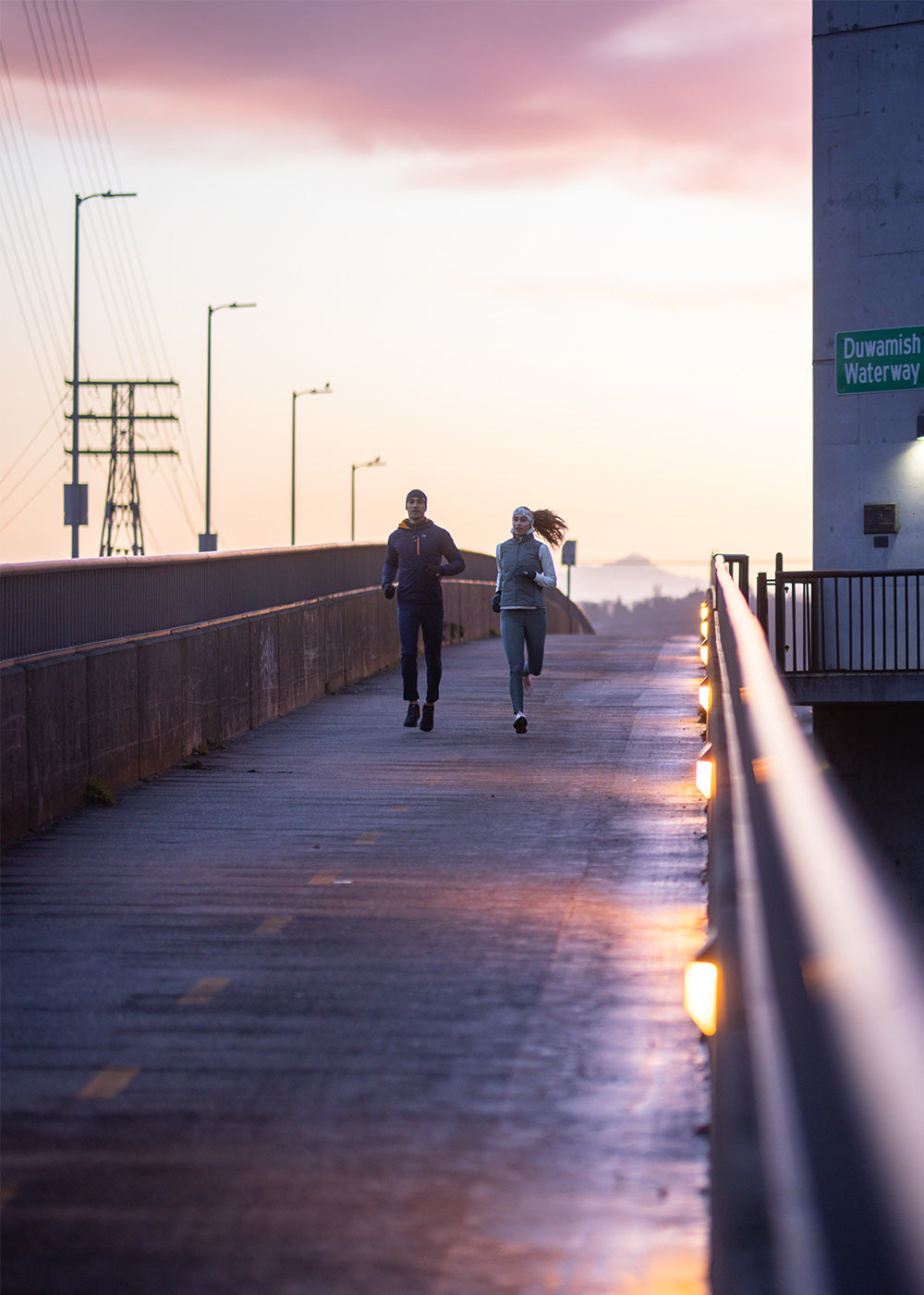 Two runners jog across a bridge along the water wearing Outdoor Research Sportwear. 