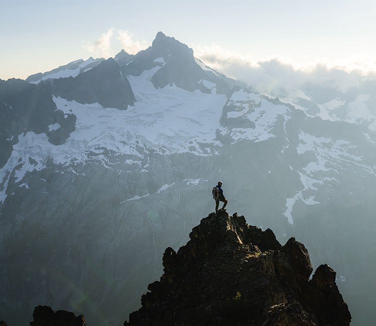 Pulled out view of a man atop a summit, with a snowcapped mountain in the background.