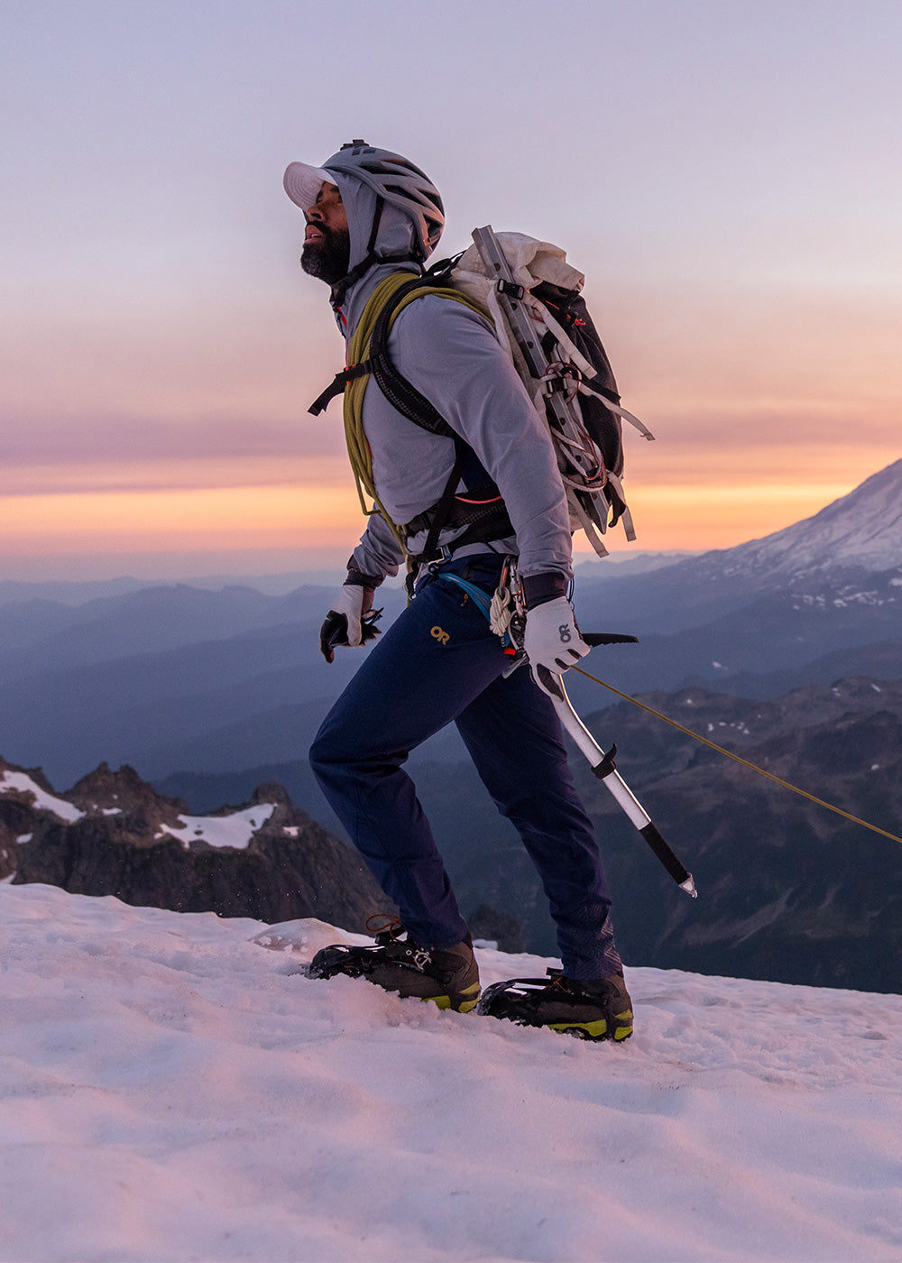 Outdoor Research athlete Max Djenohan climbs a snow-covered peak as the sun sets over the mountains in the background.