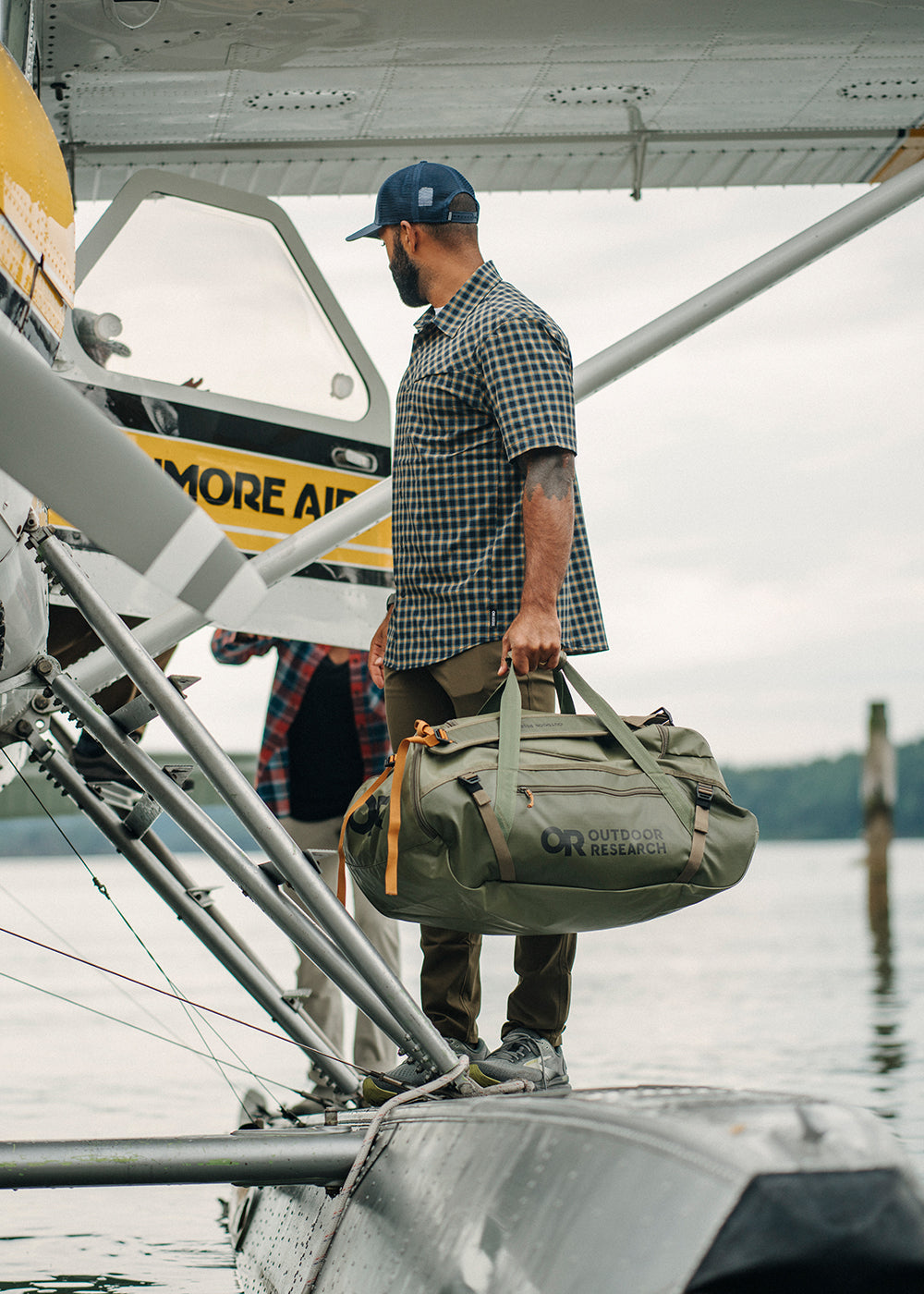 Man begins to load his Outdoor Research duffel bag onto a float plane while wearing the Astroman Short Sleeve Sun Shirt in Gold Nebula Plaid. 