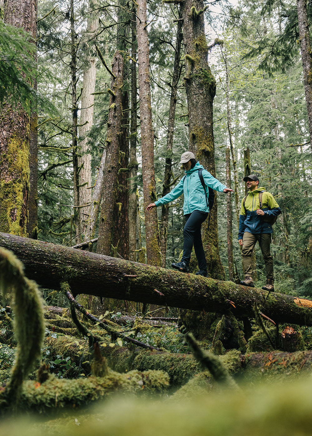 Two friends balance along a fallen down tree during their hike in the woods while wearing Outdoor Research Outerwear.