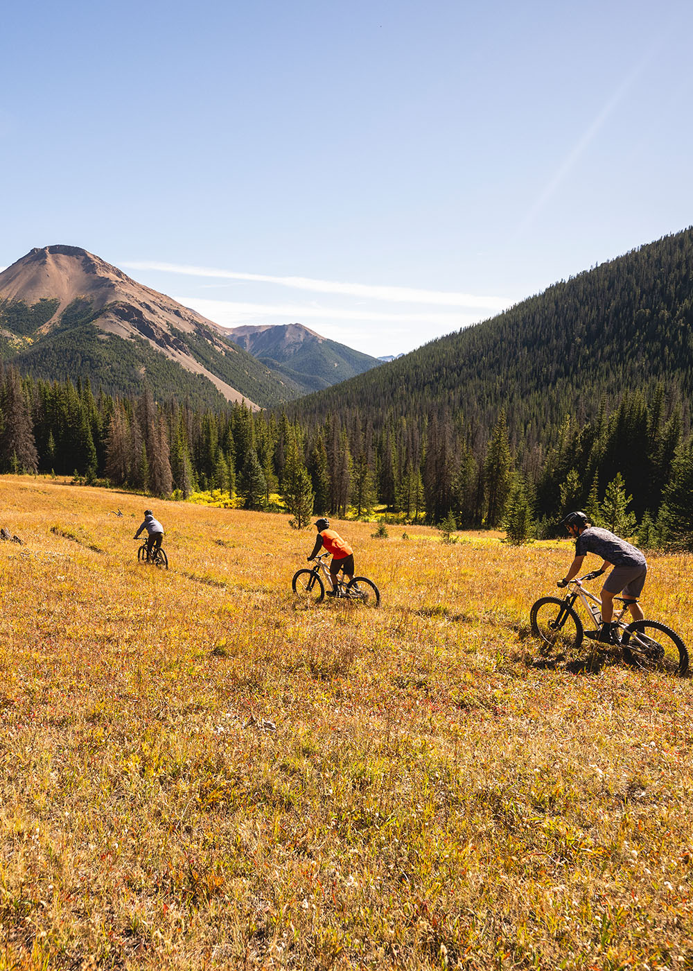 Three mountain bikers wearing Outdoor Research Sportswear ride along a dirt path beneath the blue sky. 