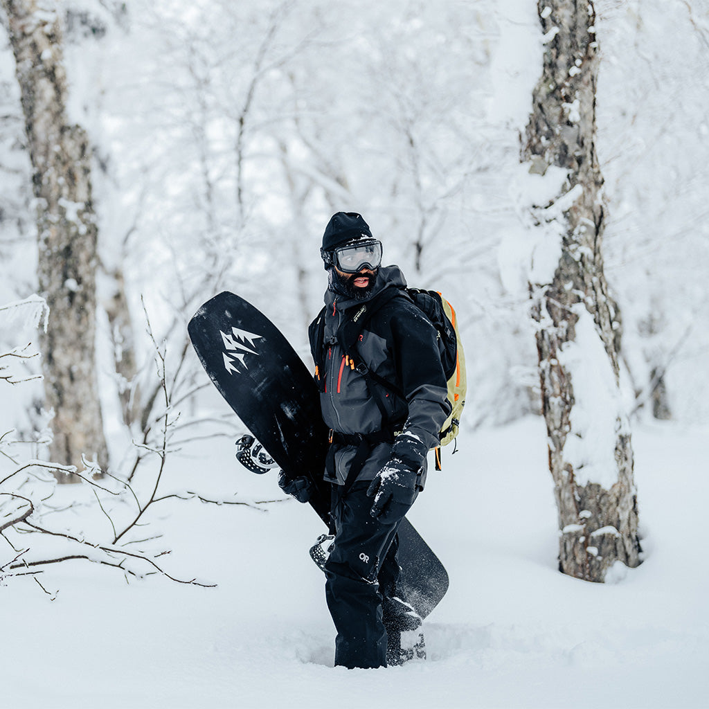 A snowboarder stays warms as he walks through the snowy forest in the Men's Headwall Gore-Tex 3L Jacket in Storm/Black.