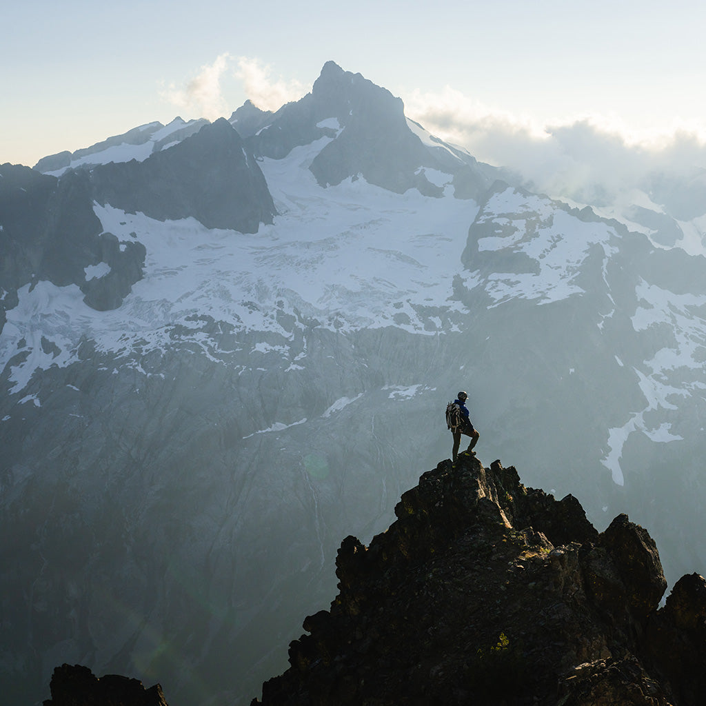 Pulled out view of a man atop a summit, with a snowcapped mountain in the background.