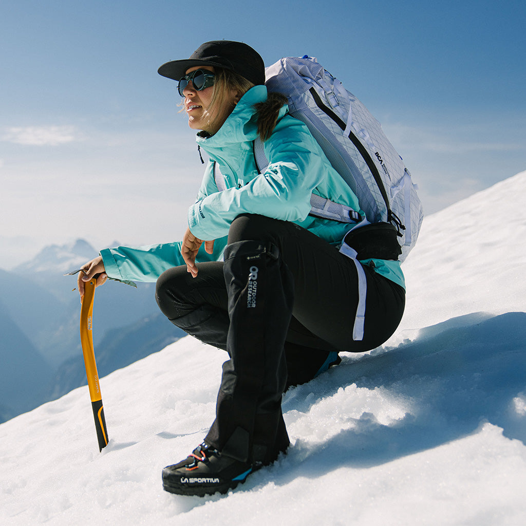 Sun beams down on a woman crouching during her hike while wearing Outdoor Research Aspire 3L Jacket in maldives/blue.